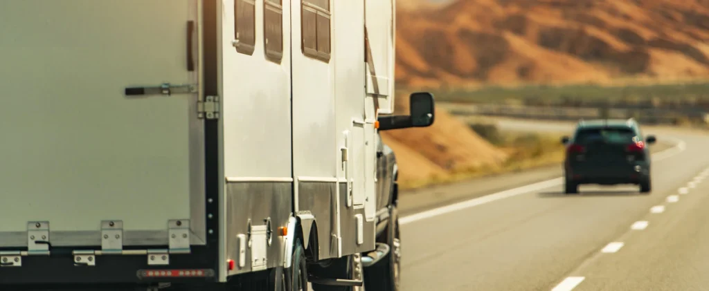 A white RV drives on a highway with mountains and desert scenery in the background, following a black car under a clear sky.