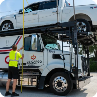 A worker in a yellow safety shirt measures the height of a white pickup truck loaded on the upper deck of a GB Cargo car carrier truck. The scene is outdoors on a sunny day.