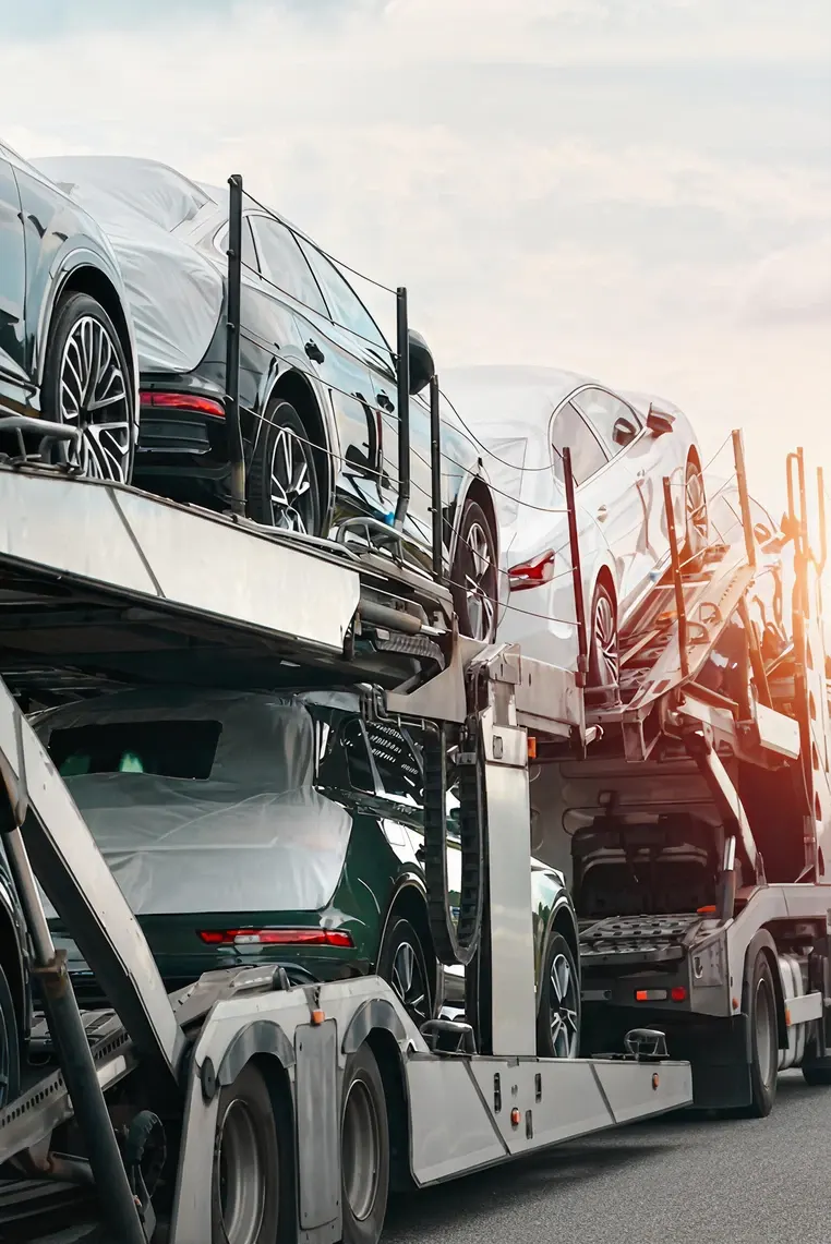 A car carrier truck transports multiple new cars, securely loaded on two levels, along a road at sunset. The vehicles are partially covered with protective wraps.
