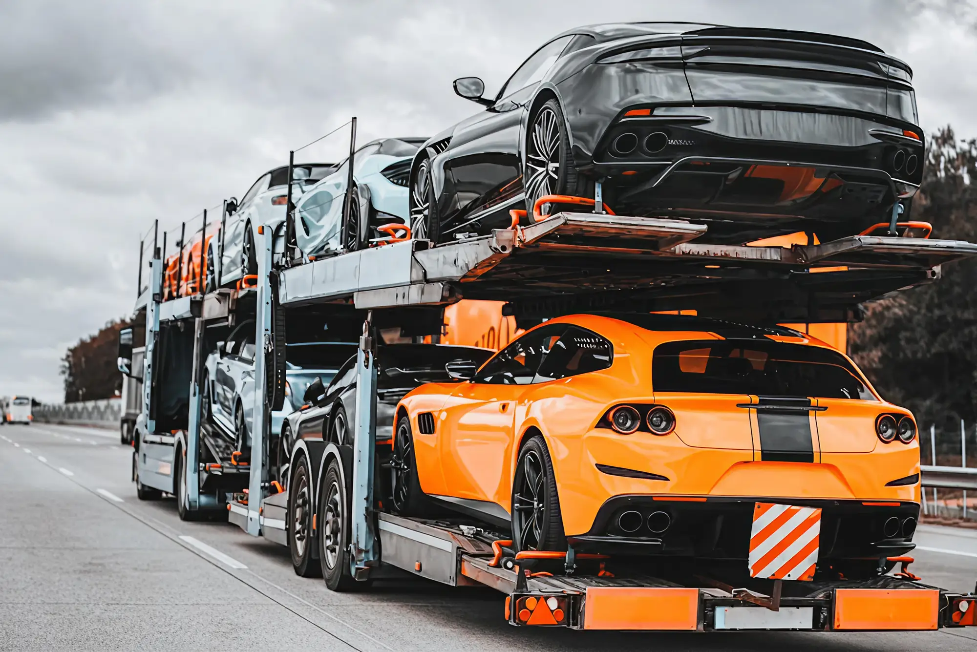 A car carrier truck transports multiple luxury sports cars, including a bright yellow car and several black cars, on a highway under a cloudy sky.