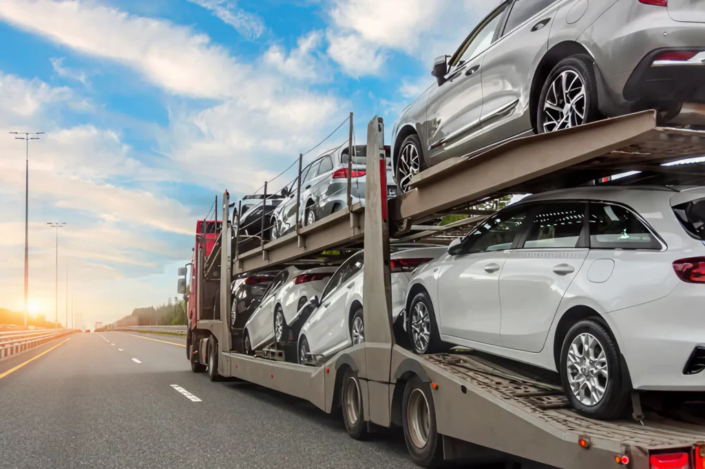 A car carrier truck loaded with new silver and white cars drives on a highway under a blue sky with clouds, heading toward the horizon at sunrise or sunset.