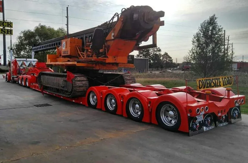 A large orange excavator is loaded onto a bright red multi-axle flatbed trailer labeled Oversize Load parked at a roadside stop. Trees and power lines are visible in the background.